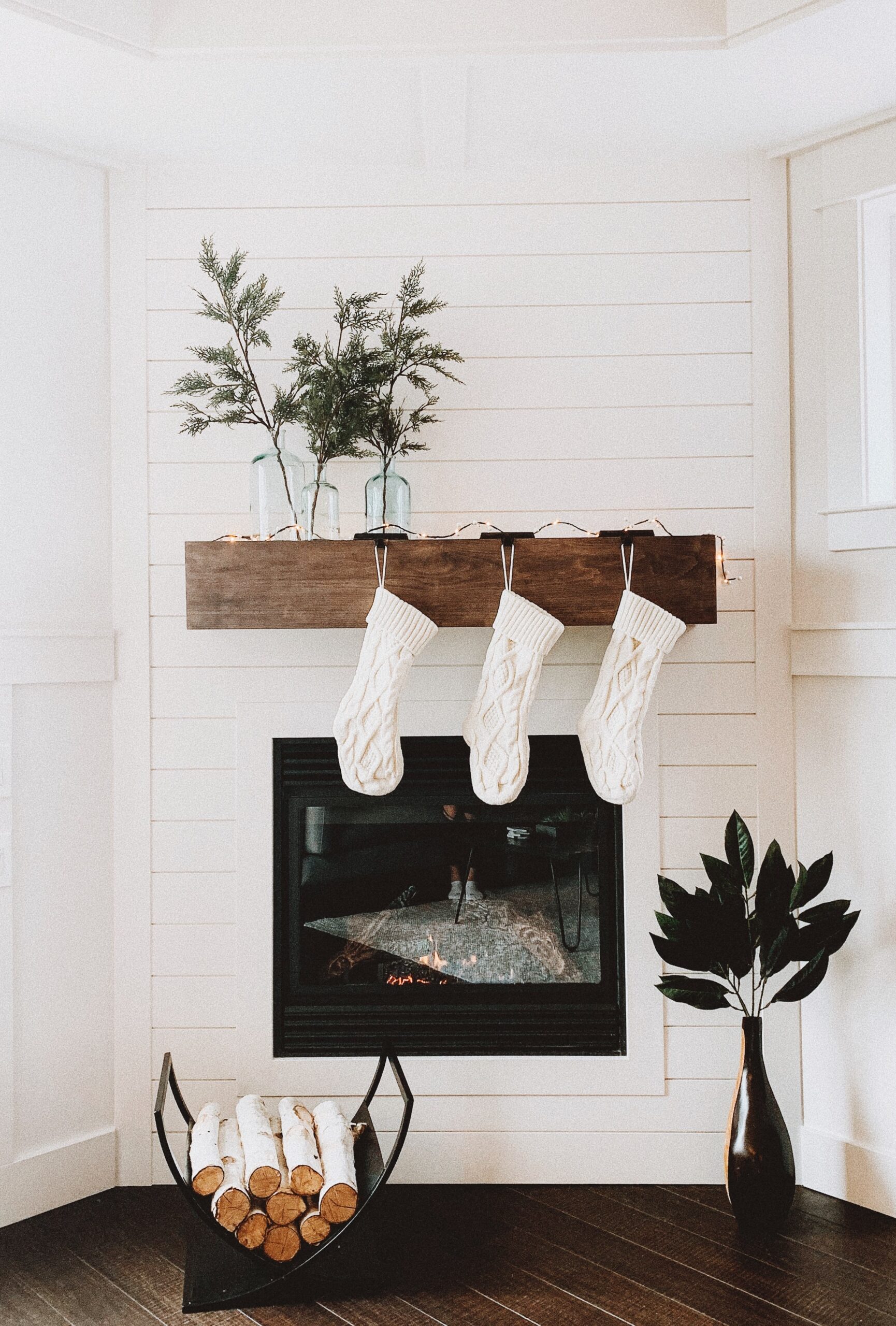 Image of a white wall featuring a wooden mantle and a black fireplace. On the mantle there is greenery and three white stockings. There is white firewood below the fireplace.