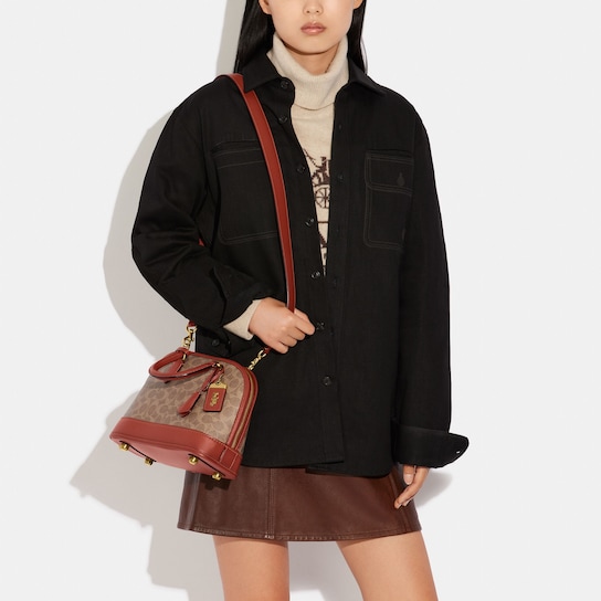 Cutoff image of a model wearing a chunky cream turtleneck, a black overshirt and a brown skirt. She is holding a brown monogrammed Coach shoulder bag with a long strap.