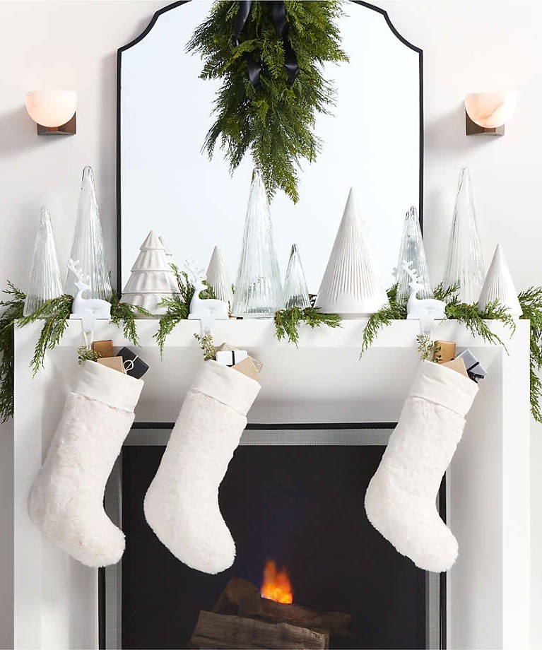 Image of a white fireplace with Christmas decor on the mantle. There is a mirror on top of the mantle, as well as various sized white Christmas trees and three white stockings. Greenery is placed throughout.