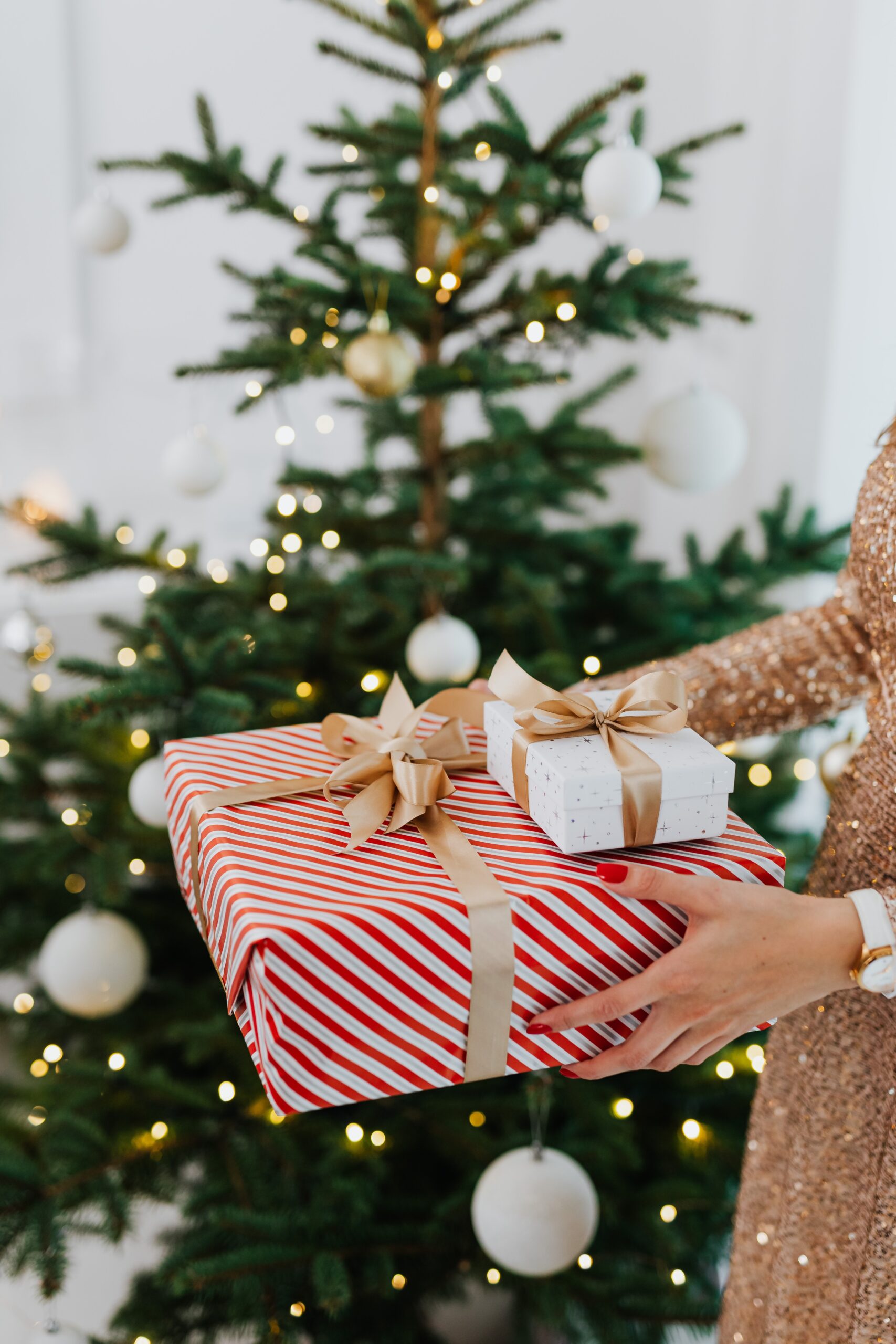 Close-up image of a woman with a red manicure holding a large gift box with red and white striped wrapping paper and a gold ribbon. A smaller white box with a white ribbon sits on top. A Christmas tree can be seen in the background.