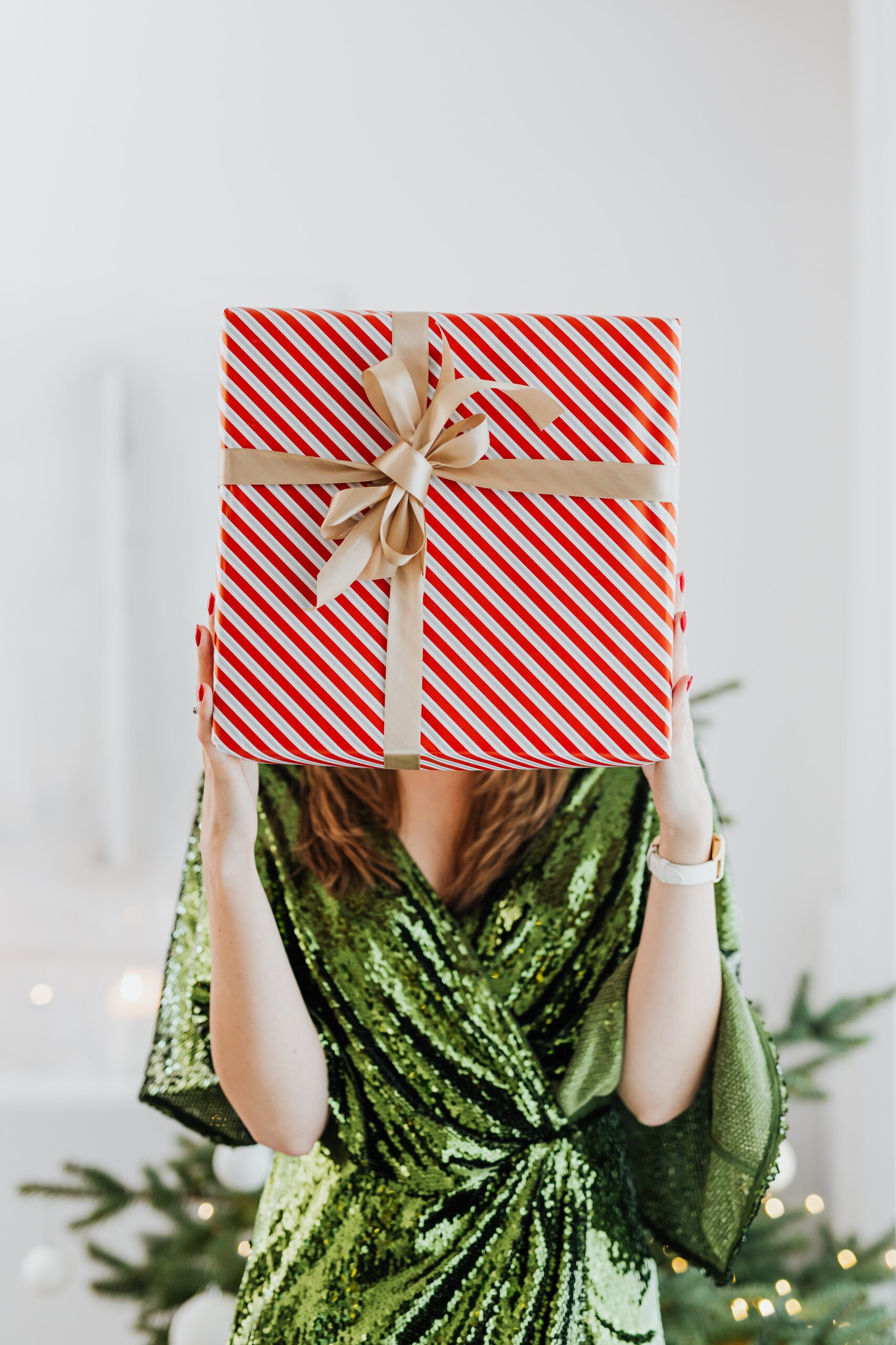 Image of a woman wearing a green sparkly dress holding a giant present wrapped in red and white wrapping paper with a gold bow in front of her face
