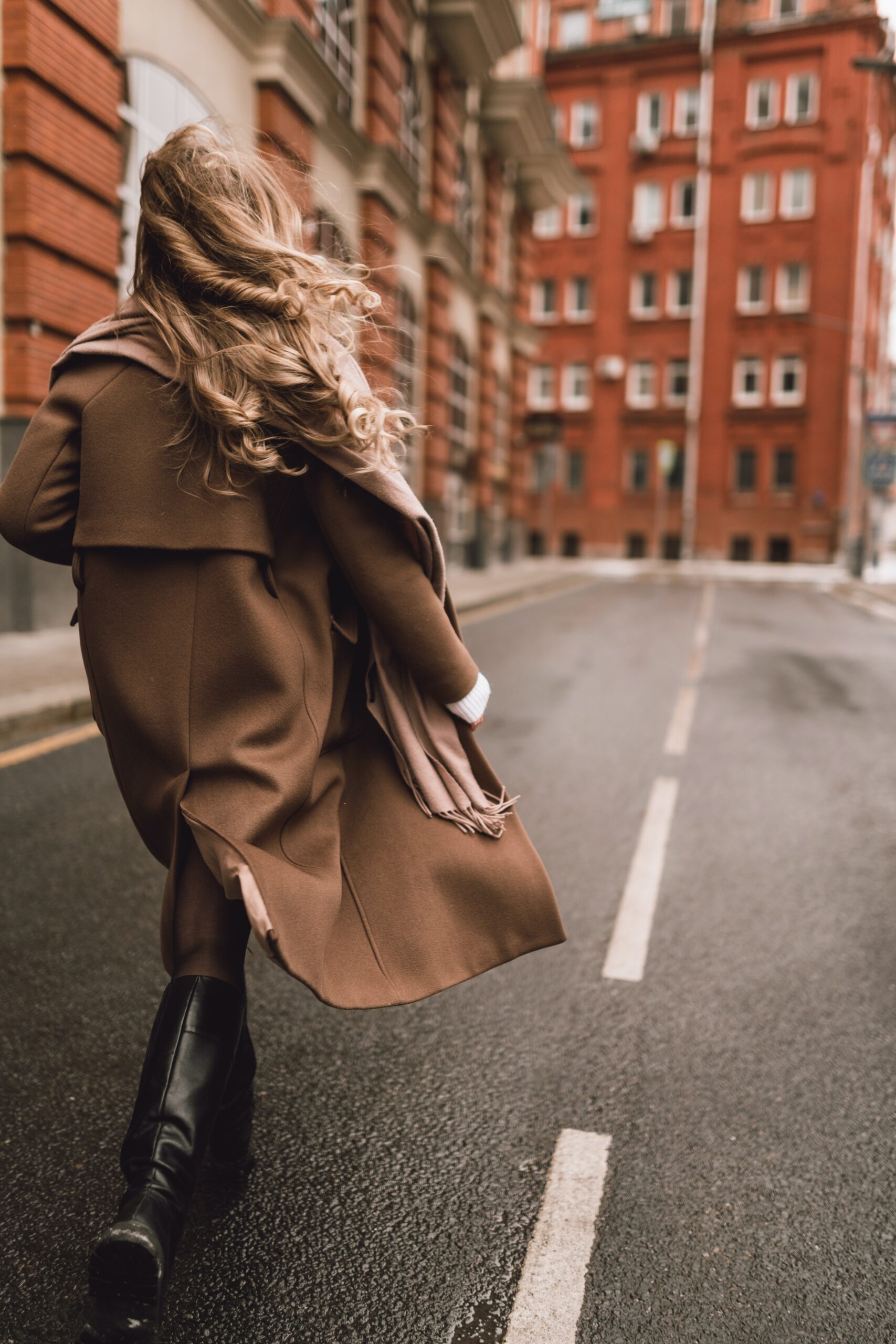 Image of the back of a woman walking down an empty street. She's wearing black knee-high boots and a brown overcoat with a cream and brown scarf and has long, dirty blonde curly hair.