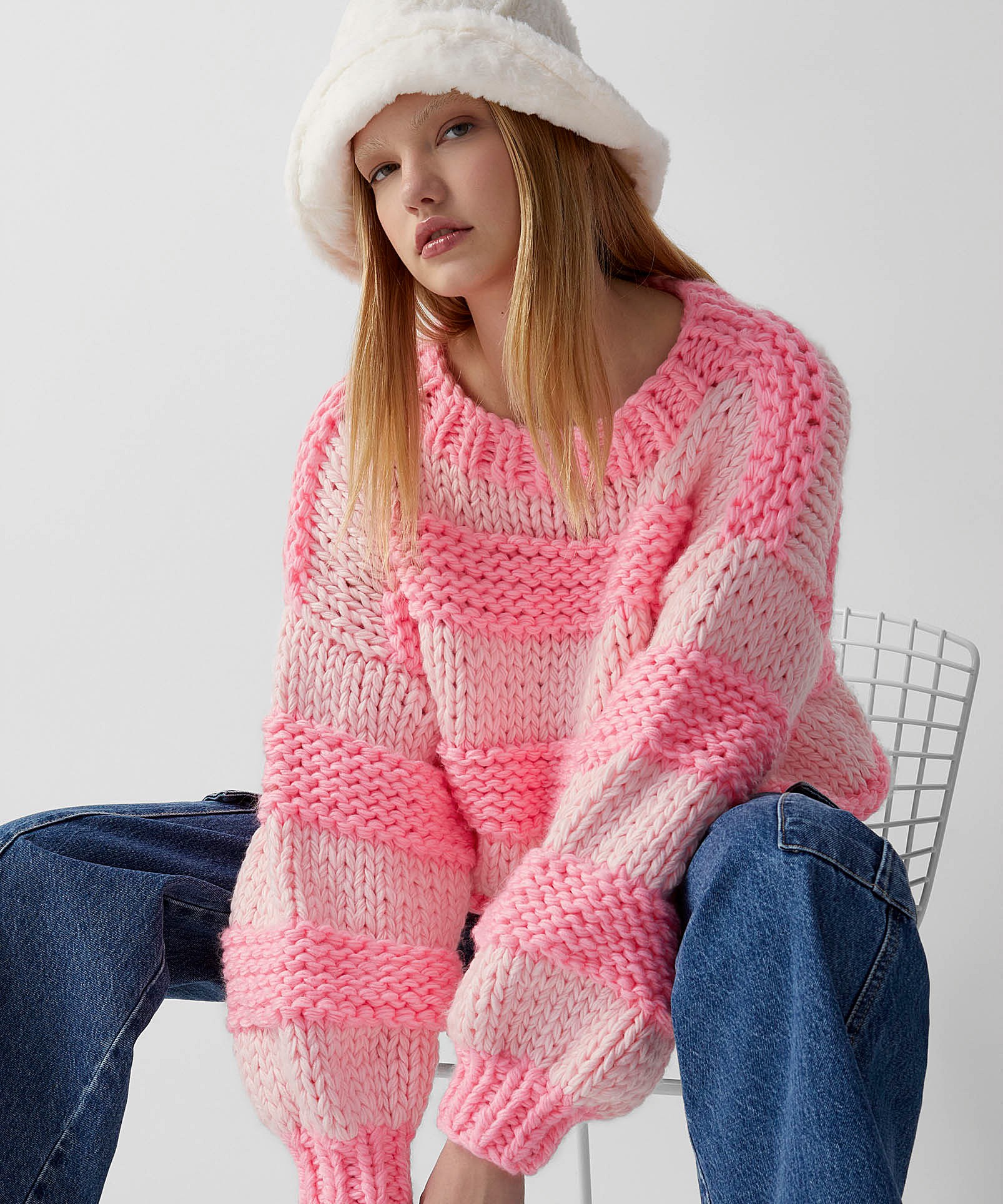 Image of a model sitting on a chair wearing a white furry bucket hat, a pink striped knit sweater and a pair of jeans.