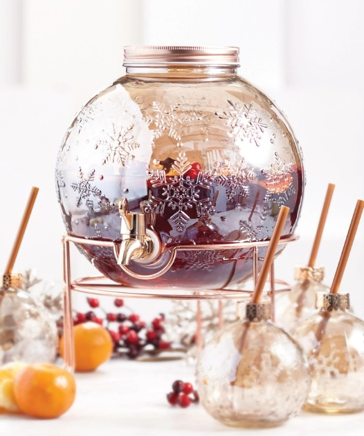 Image of a holiday-themed drink dispenser shaped like a Christmas ornament featuring a red beverage. There are four glasses (also shaped like ornaments) with straws around it, as well as some clementines and cranberries.