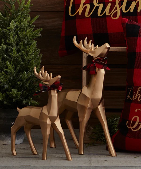 Image of bar cart decor from wyrth featuring two bronzed reindeer with red and black checkered bows around their necks and a mini Christmas tree in the background. We can see a hint of Christmas-themed pillows that are black and white checkered with gold cursive writing.