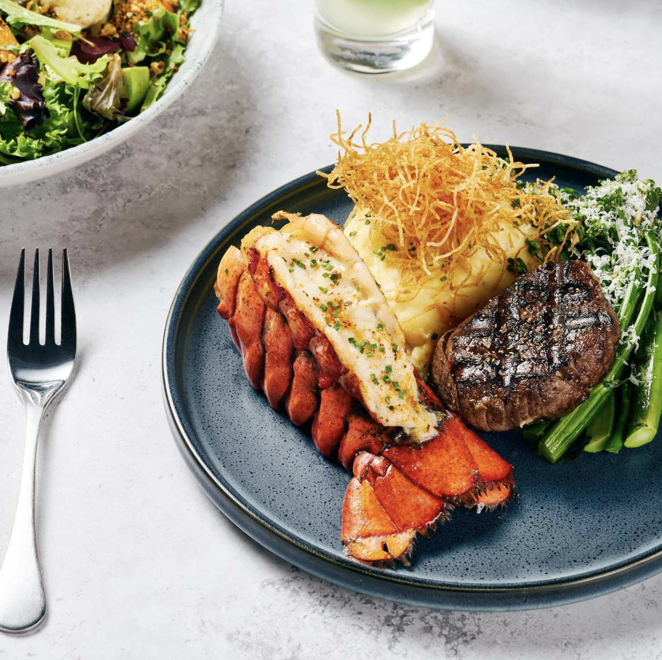 Image of a surf and turf meal at Earl's Kitchen and Bar. There is lobster tail, steak, mashed potatoes and greens on a blue plate and a bowl of salad in the left upper corner of the photo.