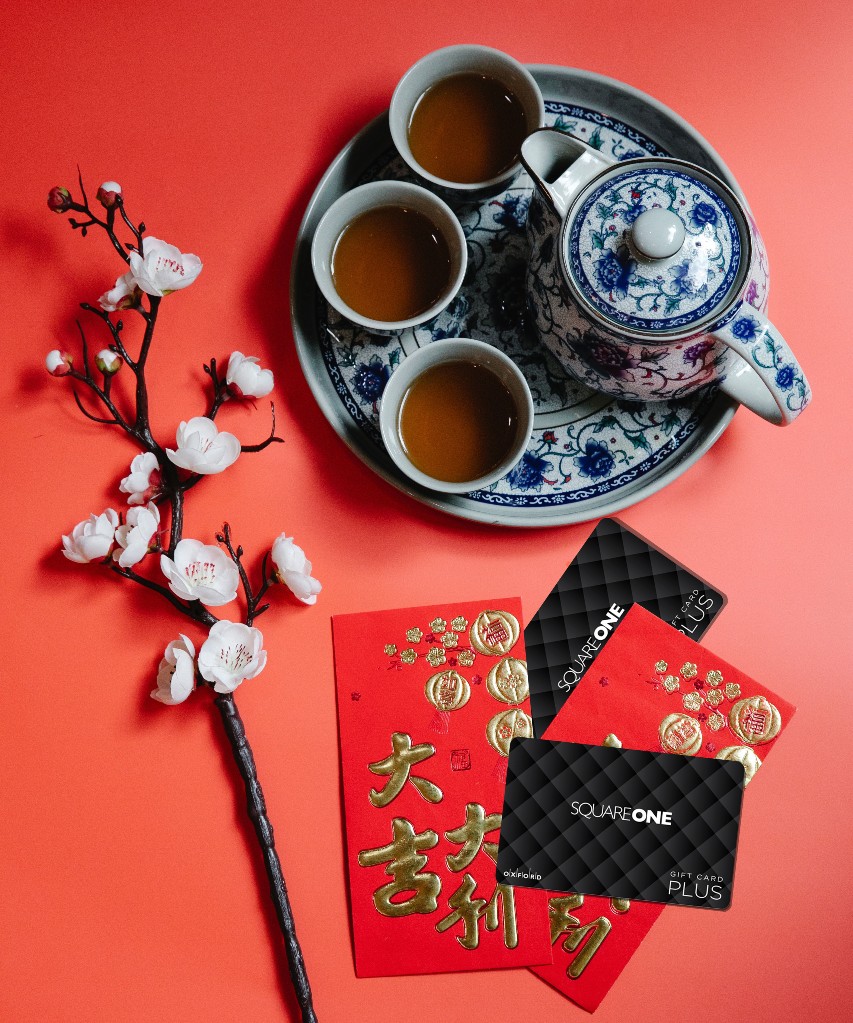 Stylized image faux cherry blossoms, a tea tray holding three cups of tea and a teapot, and Lunar New Year red envelopes with a Square One gift card layered on top. They are all placed on a red backdrop.