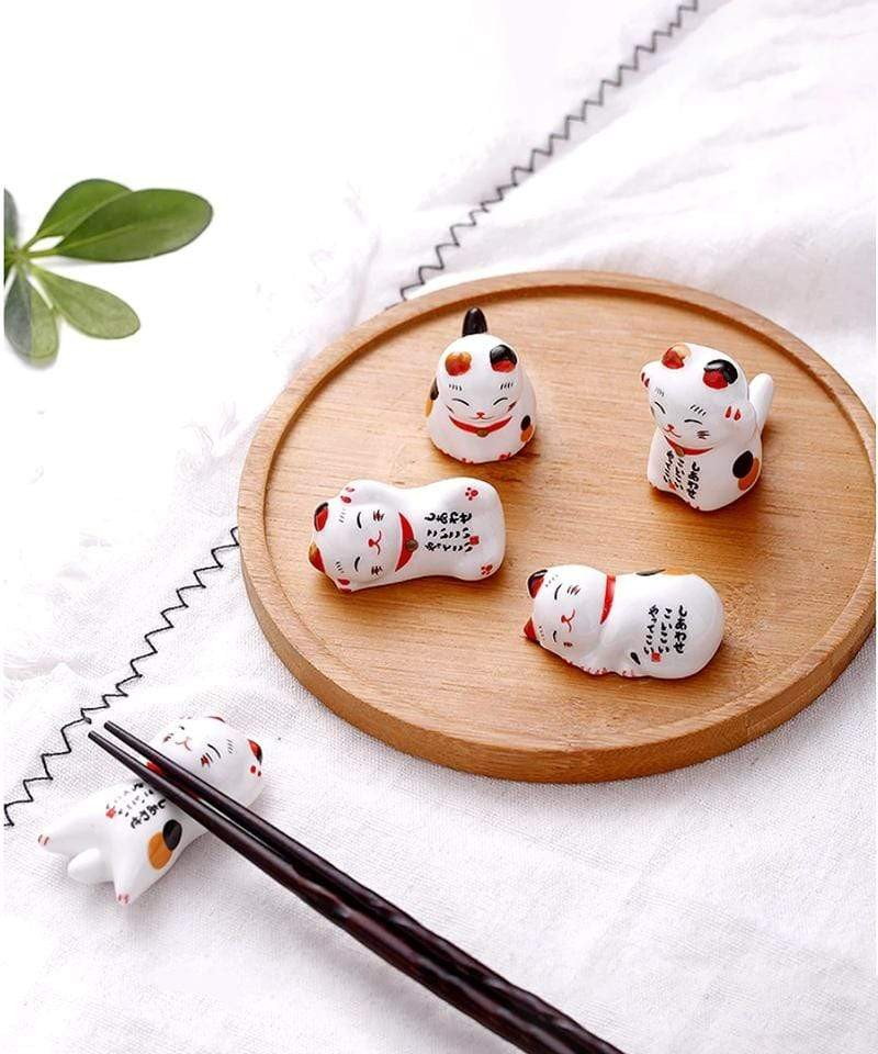 image of 5 chopstick holders in the shapes of cats in various poses. Four of the holders are on a wooden circular plate, while the fifth is on a white tablecloth holding black chopsticks.