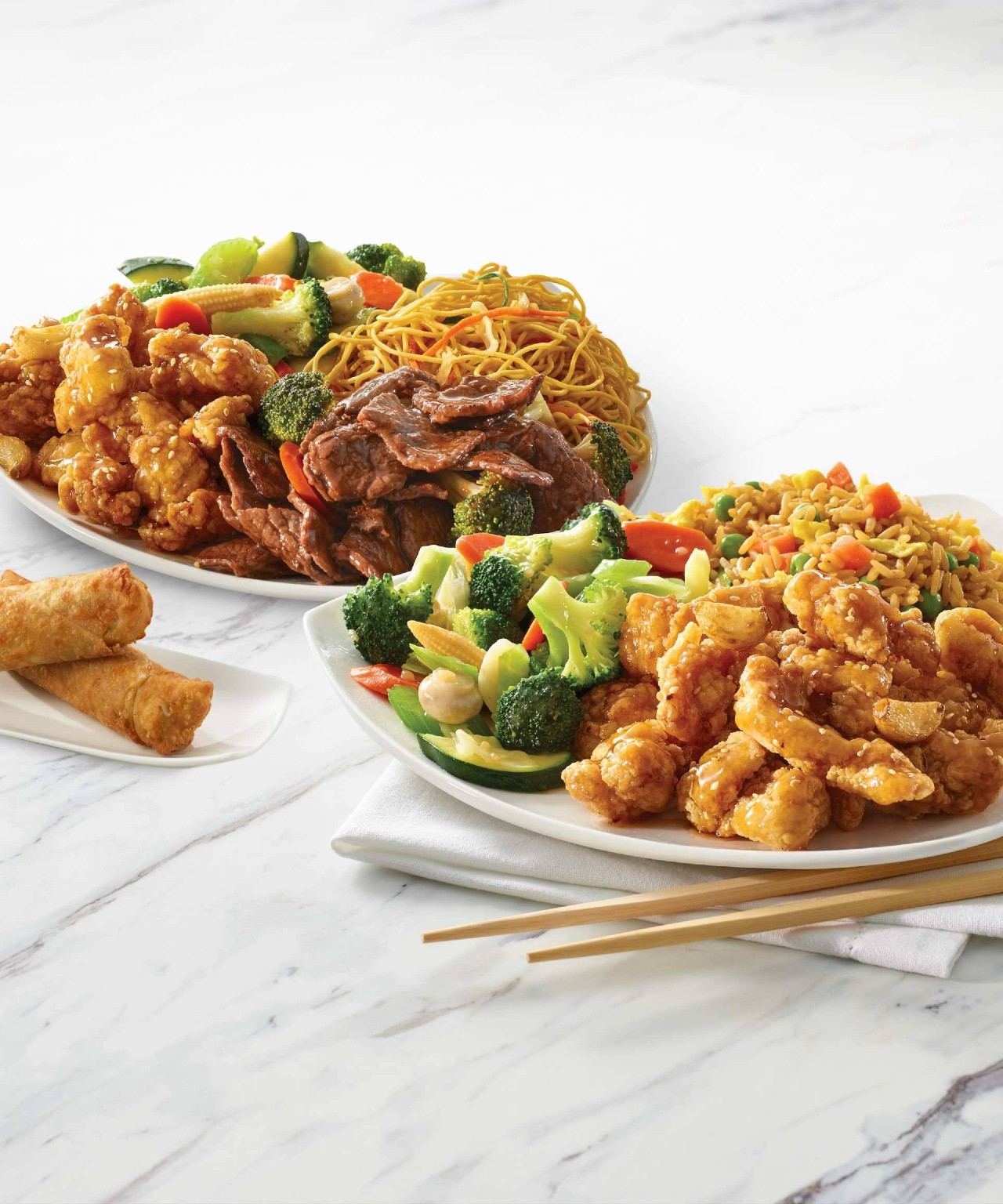 Image of two meal offerings from Manchu Wok presented on white tableware on a white marble table. One plate has noodles, fried chicken and stir fried vegetables, while the other has fried rice with vegetables and fried chicken.