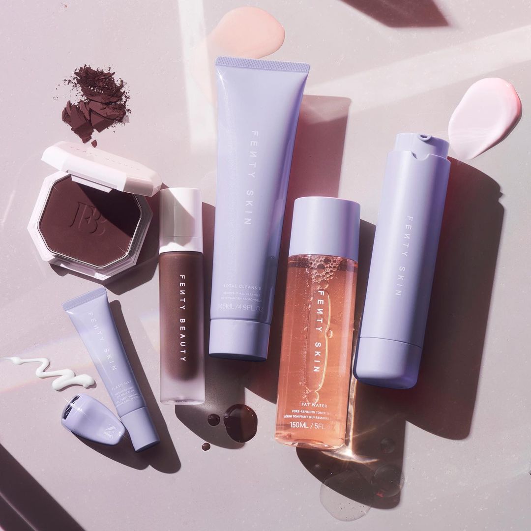 An image of six beauty and skincare products on a light purple background. There are two smaller products and three larger products. Three of the products are in purple packaging, two in clear packaging, and one is an open eyeshadow.