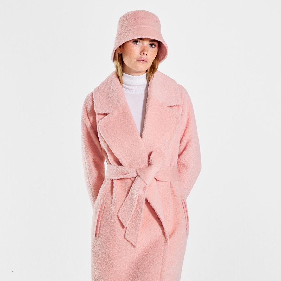 A model is standing wearing a blush pink bucket hair, a white turtleneck, and a blush pink peacoat with wide lapels and tied in a knot around her waist.