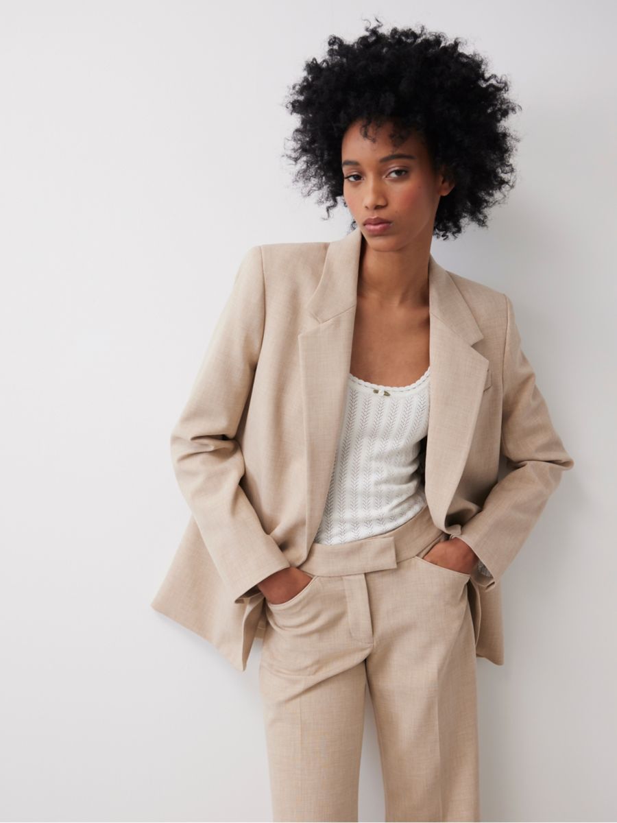 A model leaning up against a wall wearing a beige linen pant suit. The blazer is open and she is wearing a white ribbed tank top under it.