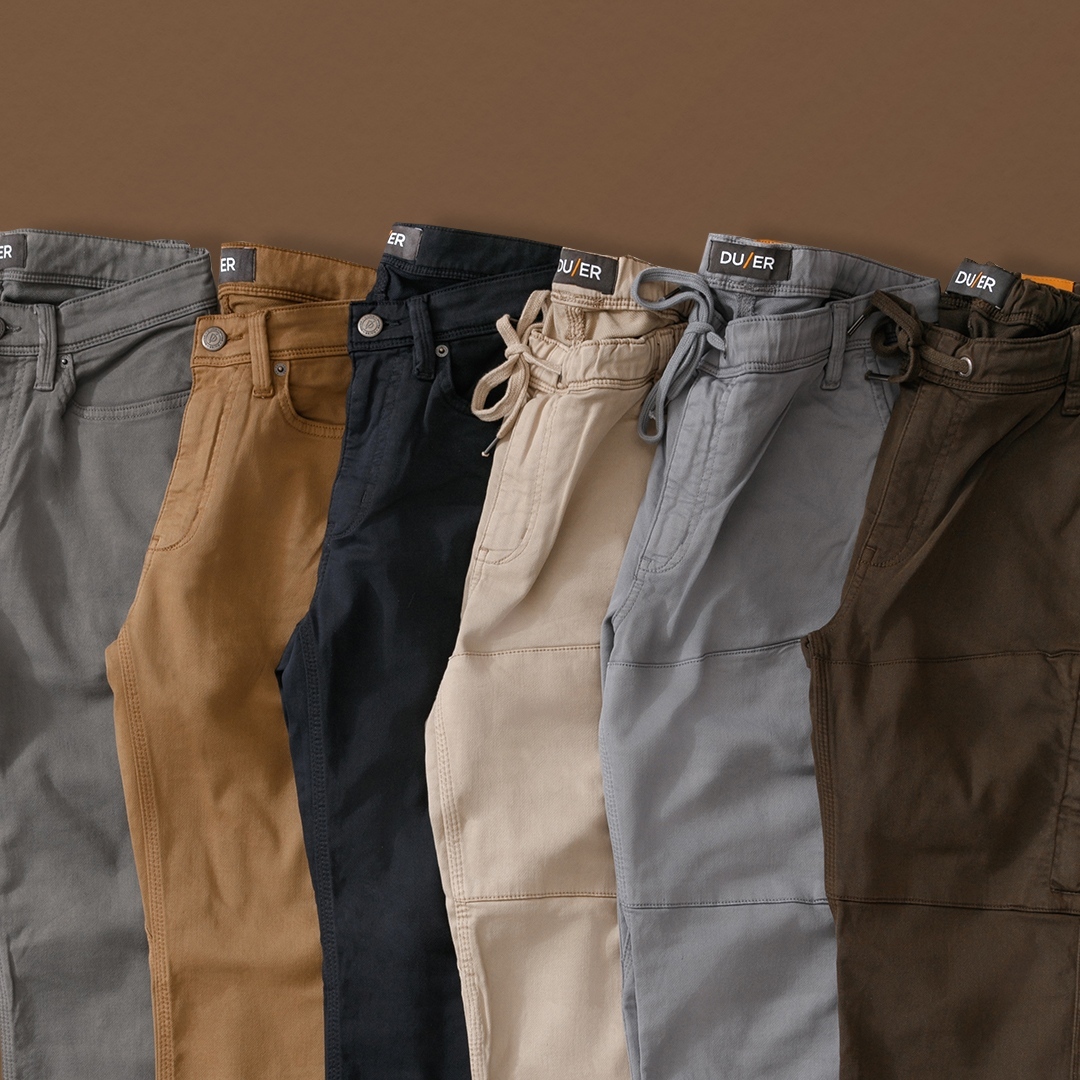 A close-up image of folded pants lined and placed in succession. From left to right the pants are dark grey, tan, black, beige, light grey and brown.