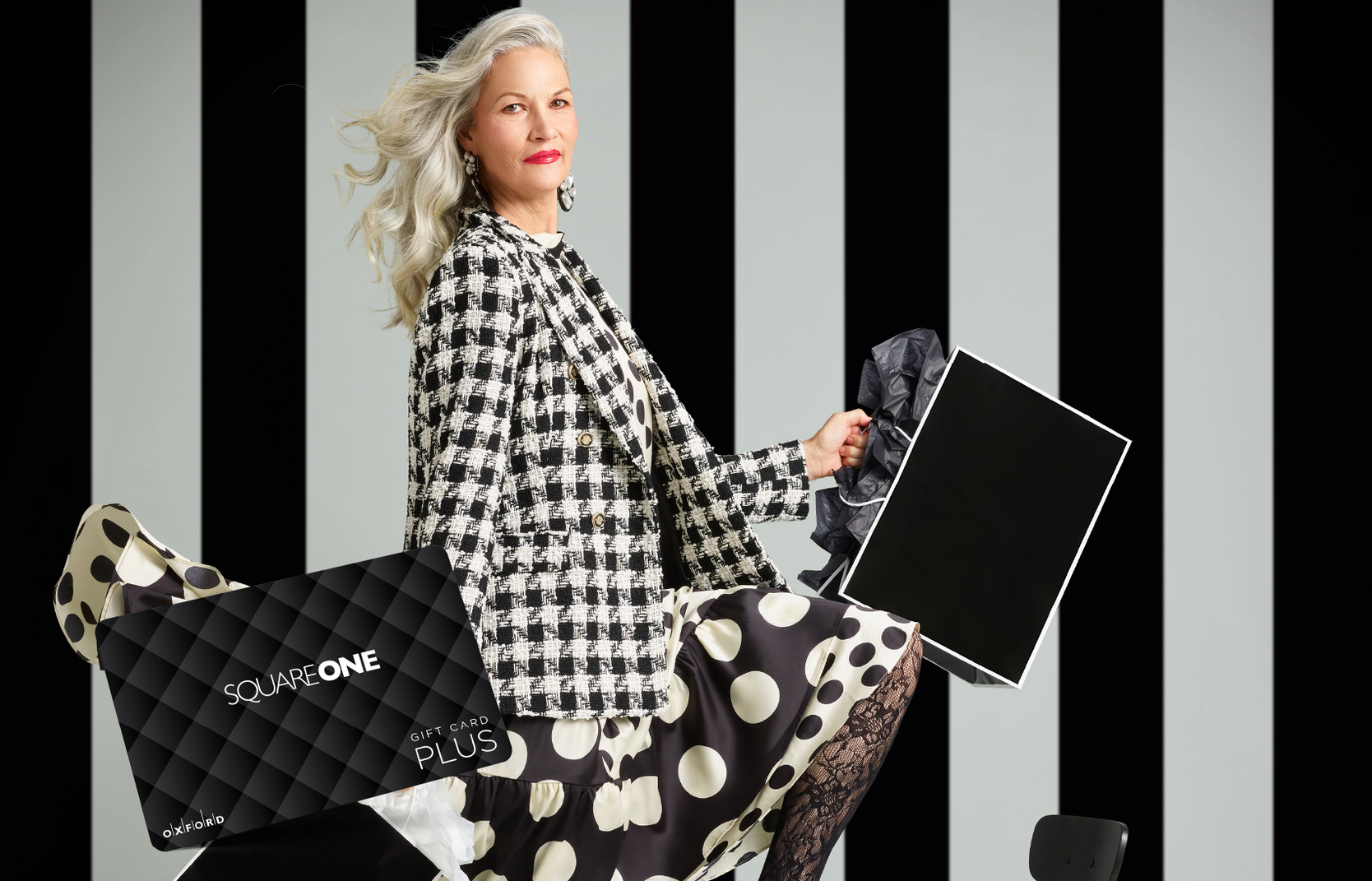 promotional image for a Square One gift card. It shows a woman wearing a black and white houndstooth blazer and black and white polka dot skirt holding black and white shopping bags. A Square One gift card is also in the image