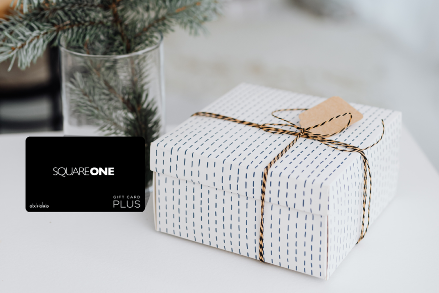 promotional image featuring a black square one gift card. beside the gift card is a present wrapped in white wrapping paper and behind the gift card is a vase with pine tree branches.
