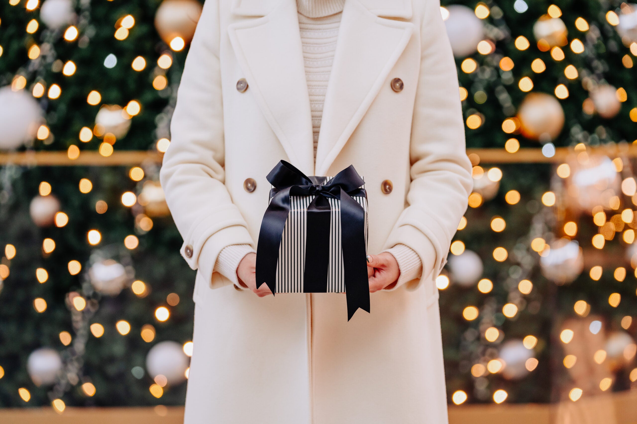 Woman with a white coat, holding a holiday grey gift with a dark bow.