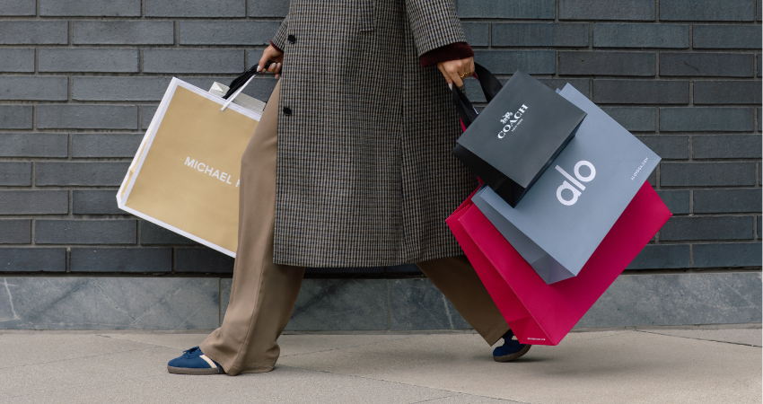 A person in a long, textured coat holding shopping bags from Michael Kors, Coach, alo, and Holt Renfrew.