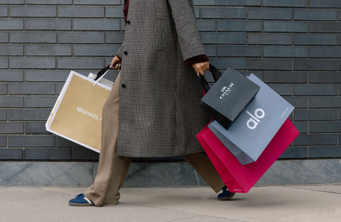 A person in a long, textured coat holding shopping bags from Michael Kors, Coach, alo, and Holt Renfrew.