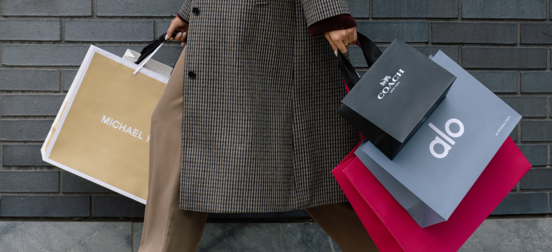 Woman with a textured brown coat holding shopping bags from Michael Kors, Coach and Alo.