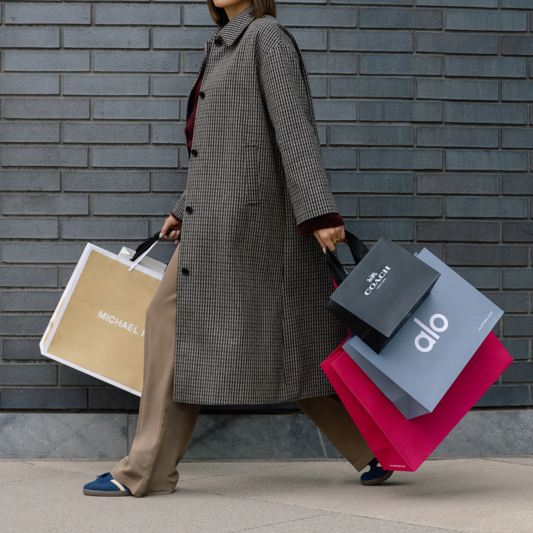 Woman with a textured brown coat holding shopping bags from Michael Kors, Coach and Alo.