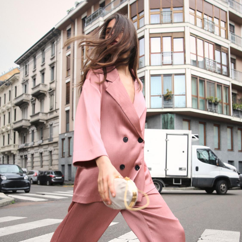 A woman wearing a silky pink double-breasted blazer is crossing the street.